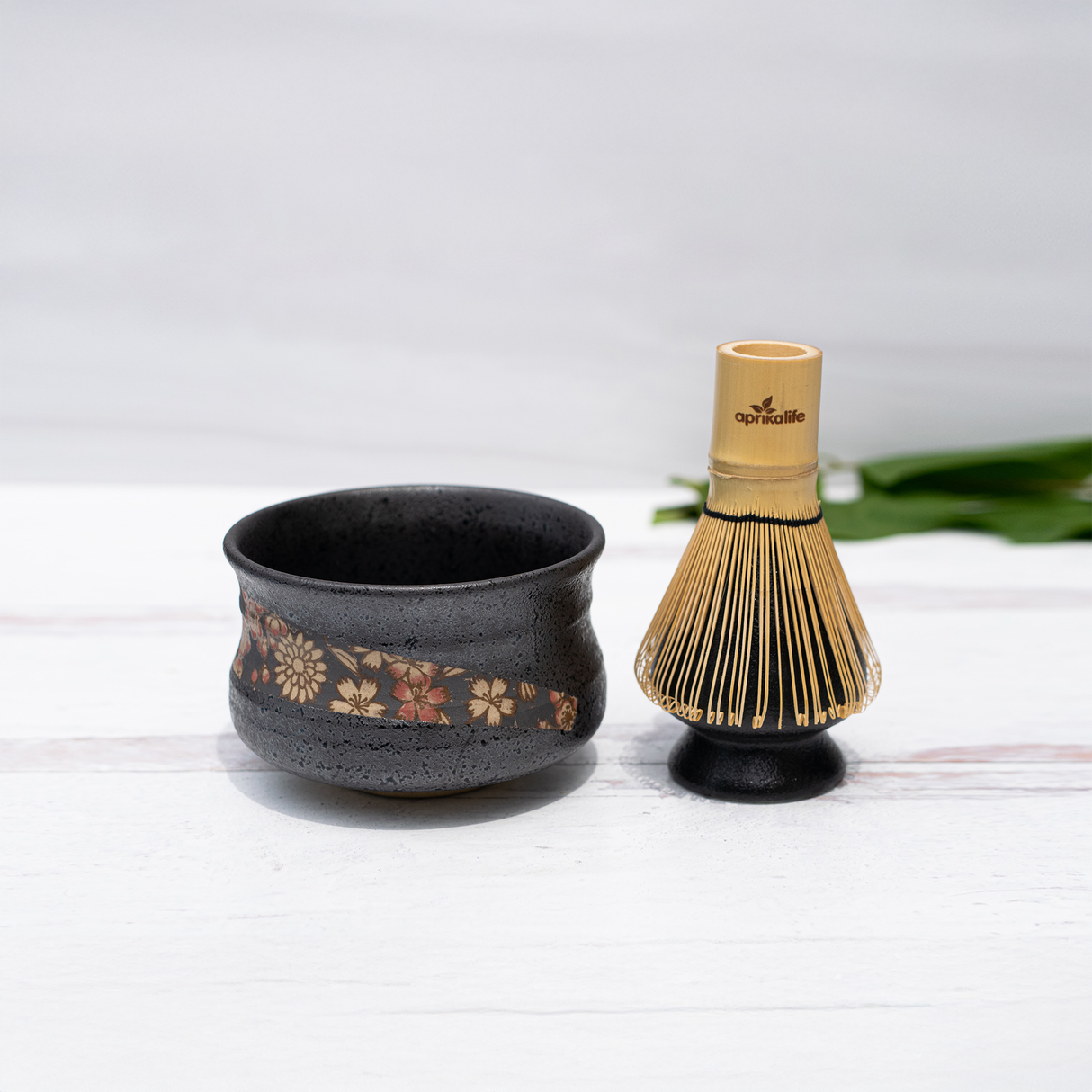 The Black with Flowers Set, featuring a black ceramic matcha bowl crafted using Minoyaki pottery techniques and adorned with a floral design, rests elegantly beside a bamboo matcha whisk on a white surface.