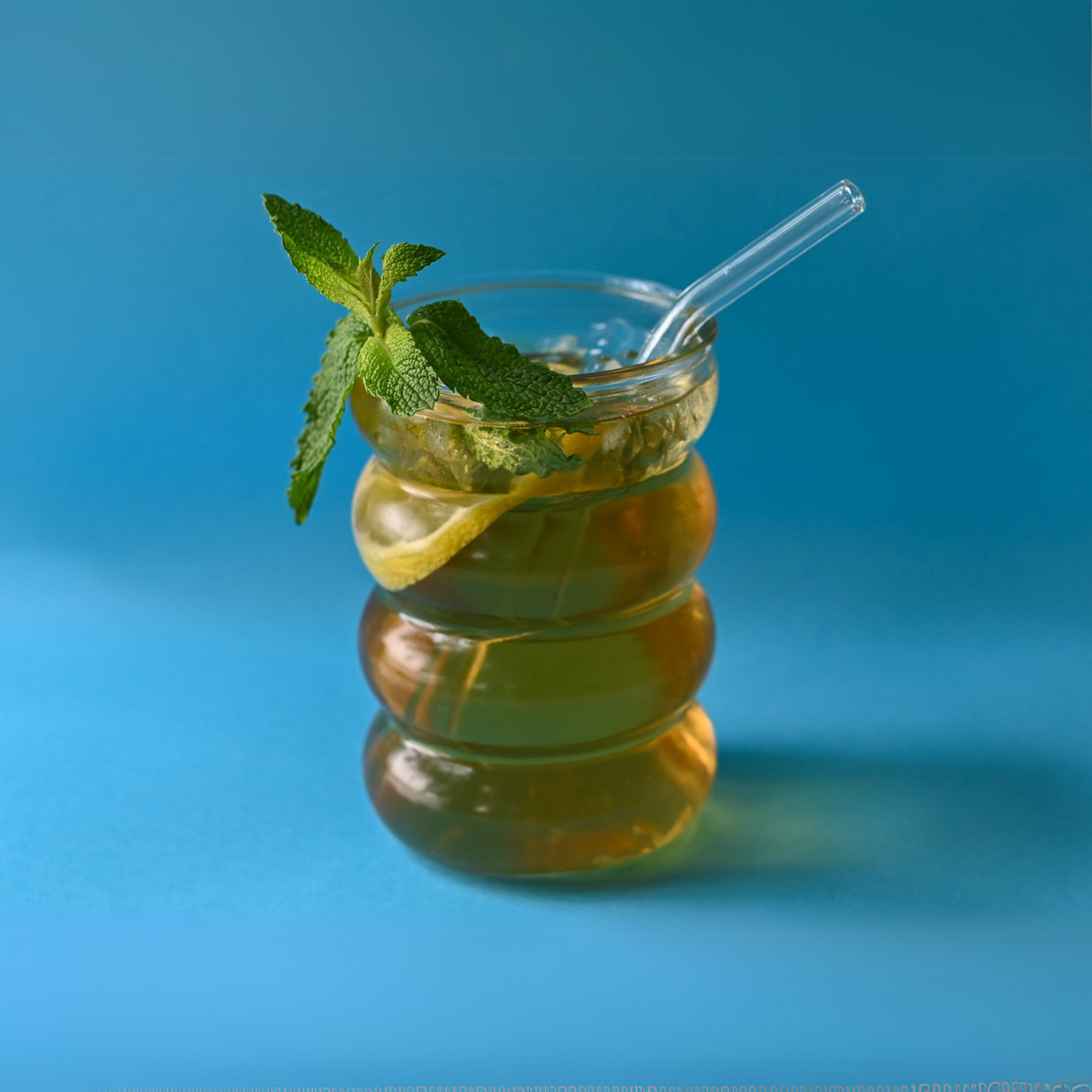 Indulge in caffeine-free 100% Pure Peppermint Herbal Tea, served iced with lemon slices, a mint garnish, and a clear straw amidst a soothing blue background. Enjoy refreshing taste and health benefits with every sip.