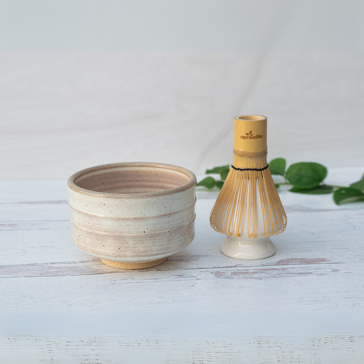 A Beige Ceramic Matcha Bowl and Bamboo Matcha Whisk from the Organic Japanese Matcha Set are elegantly displayed on a light wooden surface, framed by green leaves.