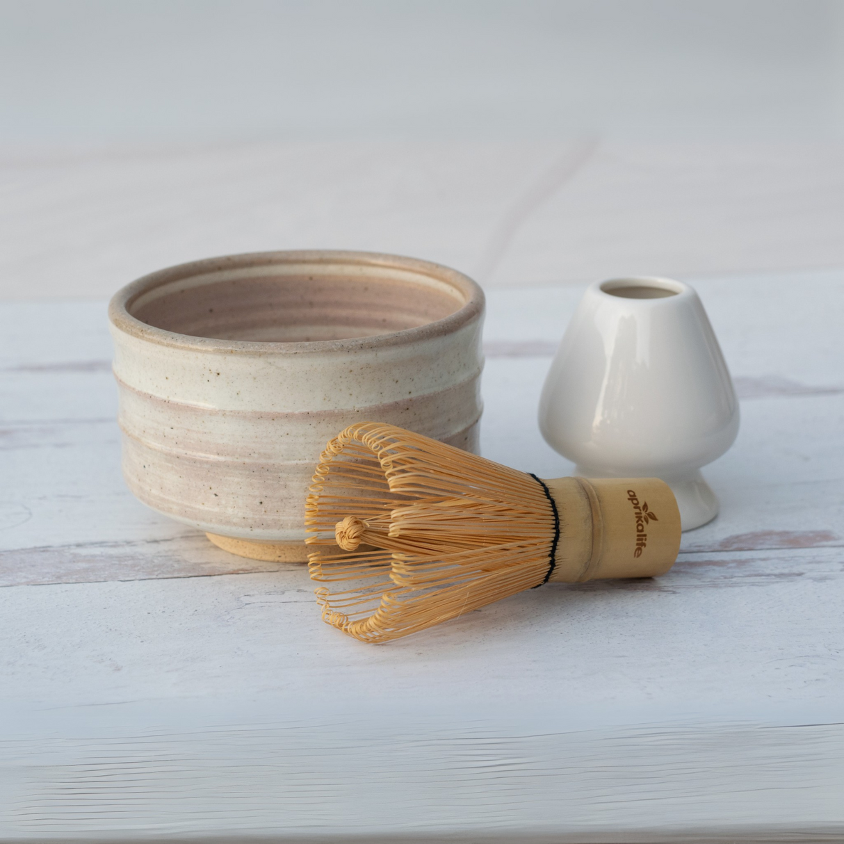 A Beige Ceramic Matcha Bowl, Bamboo Matcha Whisk, and Whisk Holder set on a white wooden surface exemplify elegant matcha accessories crafted from Minoyaki pottery.
