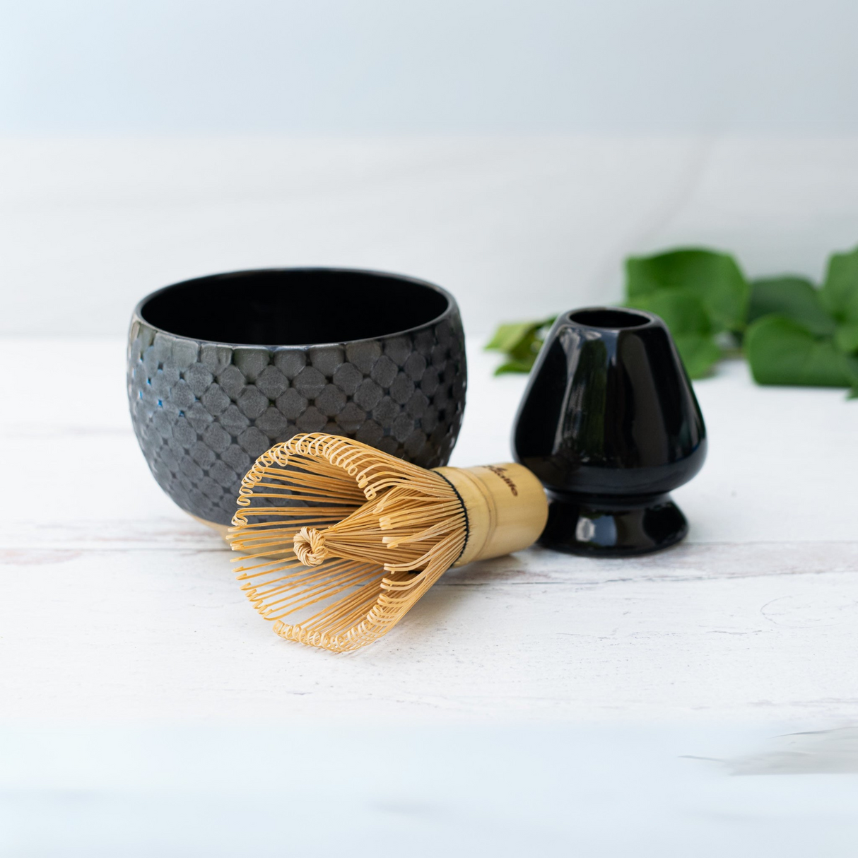 The Black Ami Pattern Set includes a bamboo whisk, a black ceramic matcha bowl, and a whisk holder, all arranged on a light surface with lush green leaves for an elegant touch.