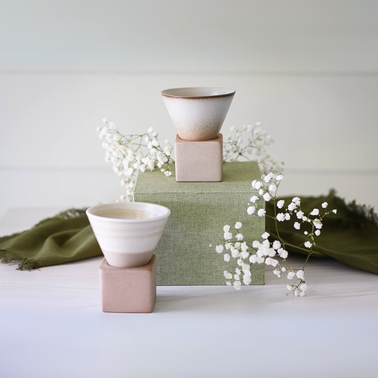 Two Brown Coffee Mug with Base - 6.8 oz/200ml sit elegantly on blocks, framed by green fabric and delicate white flowers, showcasing exquisite ceramic artistry.