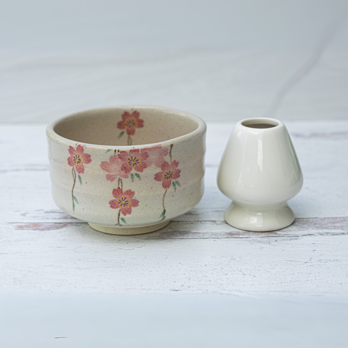 The Pink Sakura Flowers Ceramic Matcha Bowl, Bamboo Whisk, Whisk Holder, and Organic Japanese Matcha Set features a floral ceramic bowl paired with a white whisk holder—ideal for fans of Japanese pottery and authentic matcha teaware.