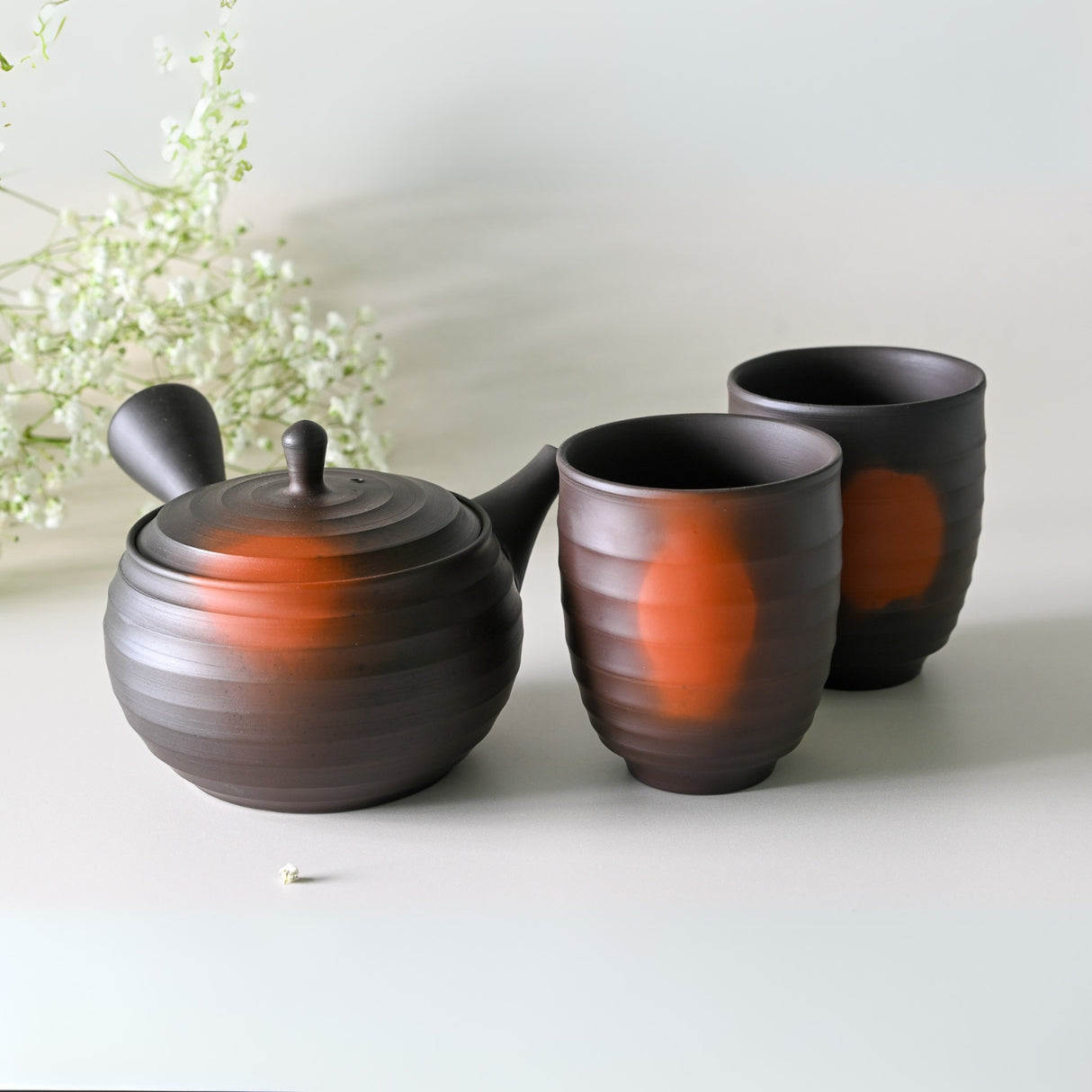 The Japanese Kyusu Set, featuring a 270ml teapot with filters and two matching cups with orange accents, rests elegantly on a light surface adorned with green foliage.