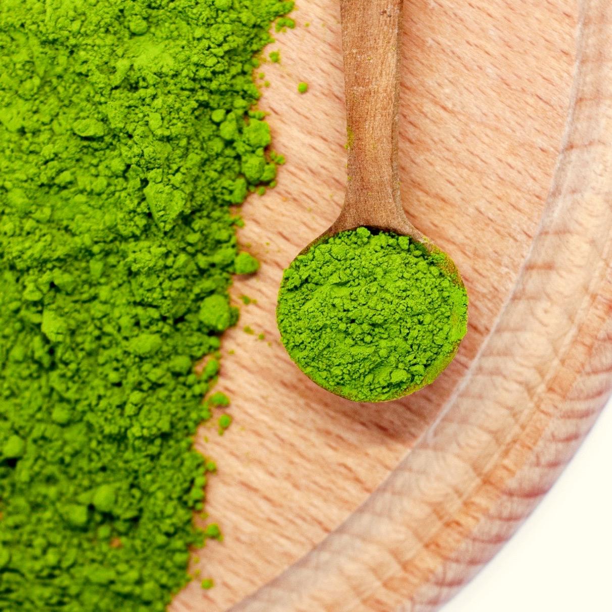 A wooden spoon with Organic Japanese Matcha Green Tea Powder rests on a wooden surface, surrounded by vibrant green matcha powder.