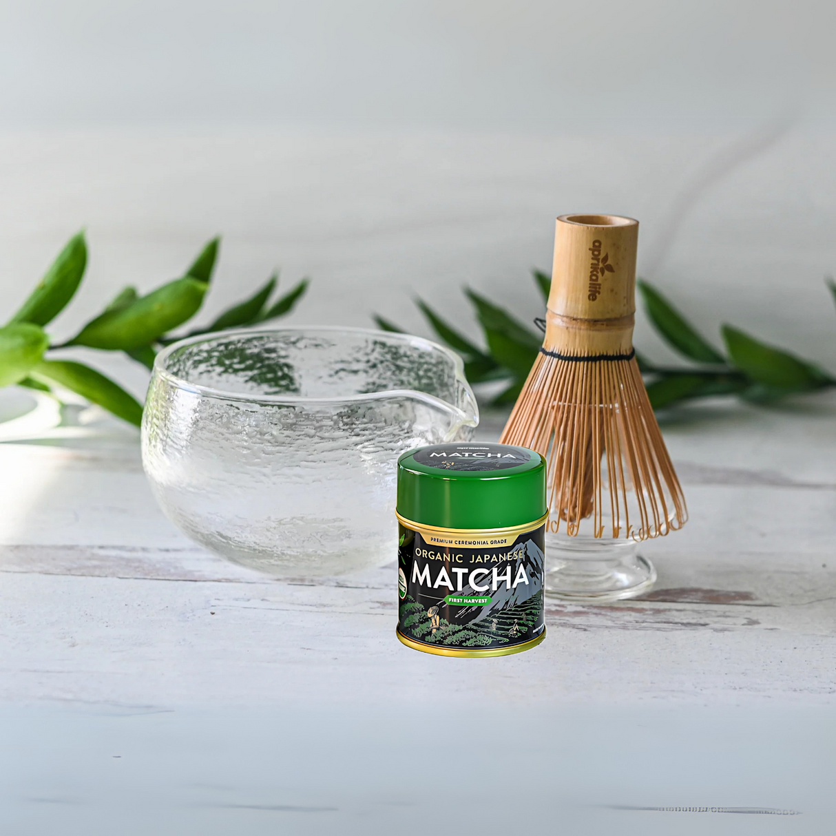 The Textured Spouted Glass Matcha Bowl Pattern 1, Bamboo Matcha Whisk, Whisk Holder, and Organic Japanese Matcha Set are displayed on a white surface with green leaves.