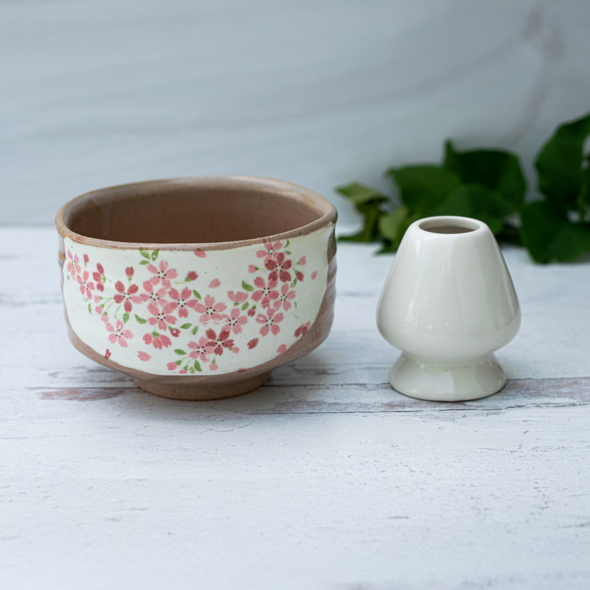 The Big Sakura Flowers Matcha Bowl, Bamboo Matcha Whisk, Whisk Holder, and Organic Japanese Matcha Set are displayed on a light wooden surface with green leaves in the background, highlighting the refined beauty of Japanese tea culture.
