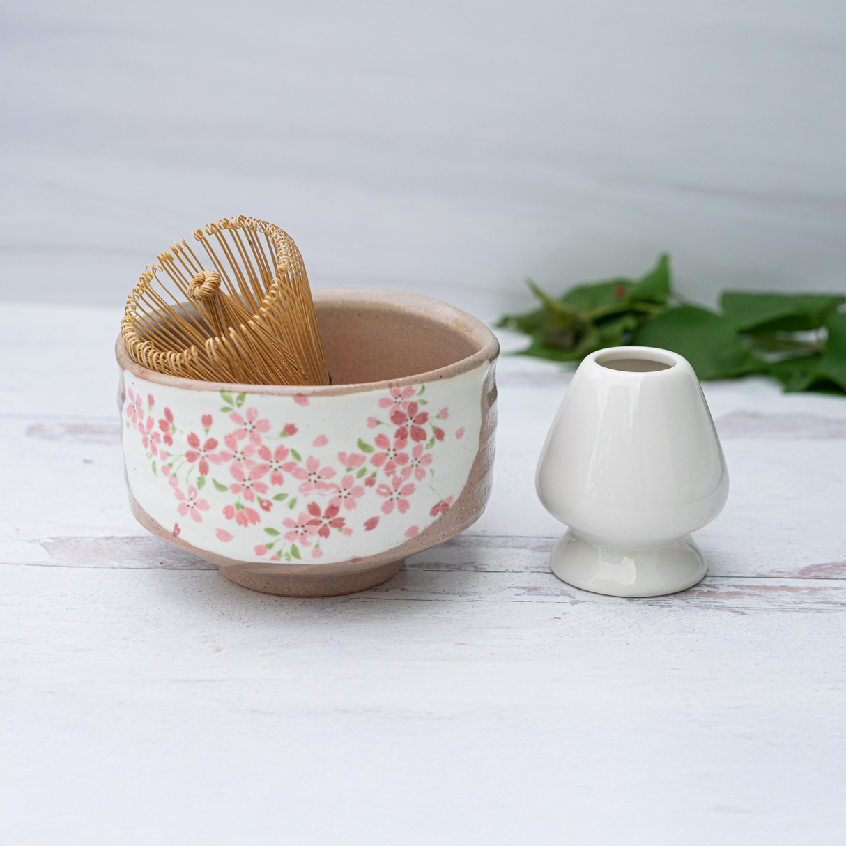 The Big Sakura Flowers Matcha Bowl, Bamboo Matcha Whisk, Whisk Holder, and Organic Japanese Matcha Set are displayed on a light surface with green leaves in the background.