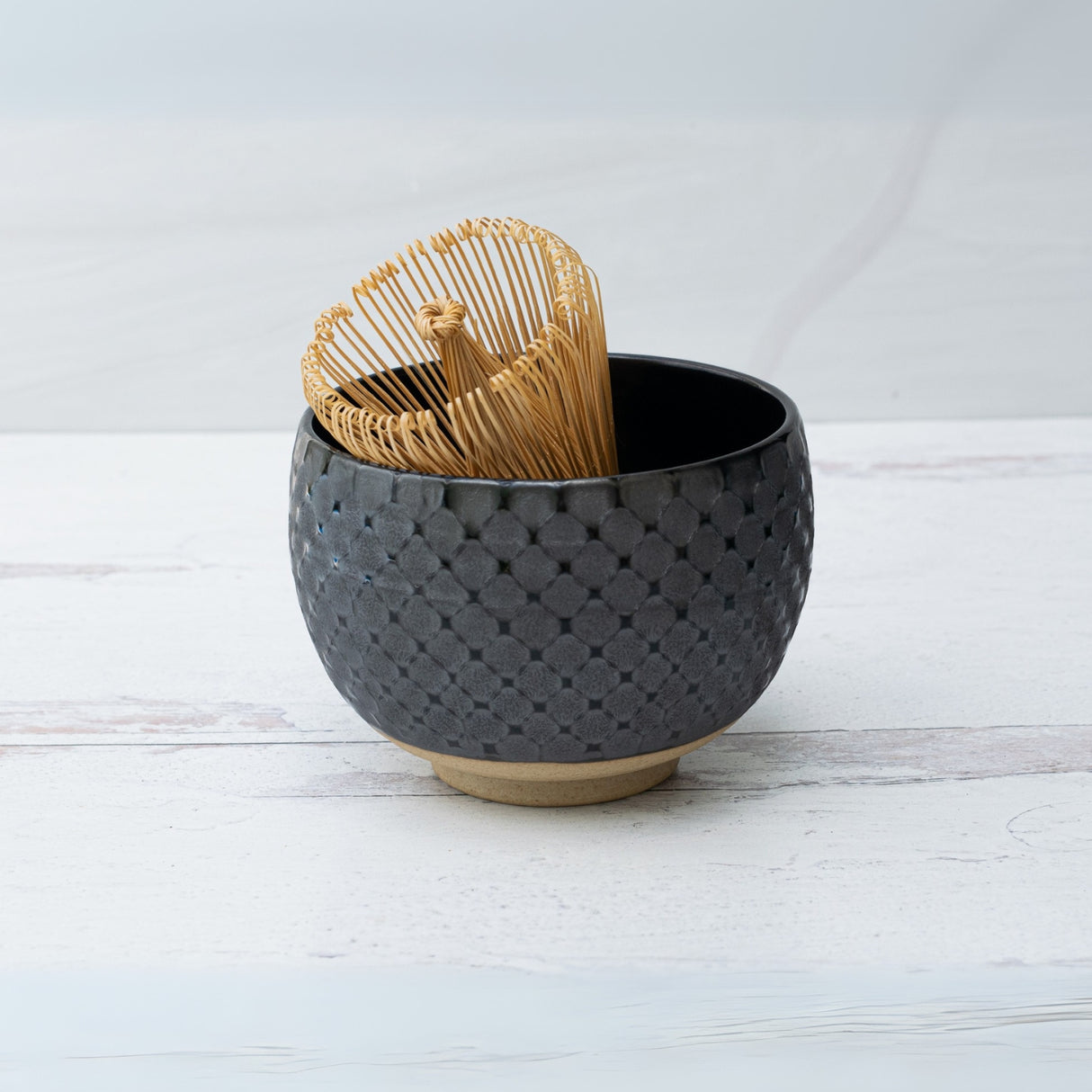 Highlighted by the bamboo whisk inside, the Black Ami Pattern Matcha Bowl, featuring a Japanese pottery style with a textured finish, rests on a light wooden surface.