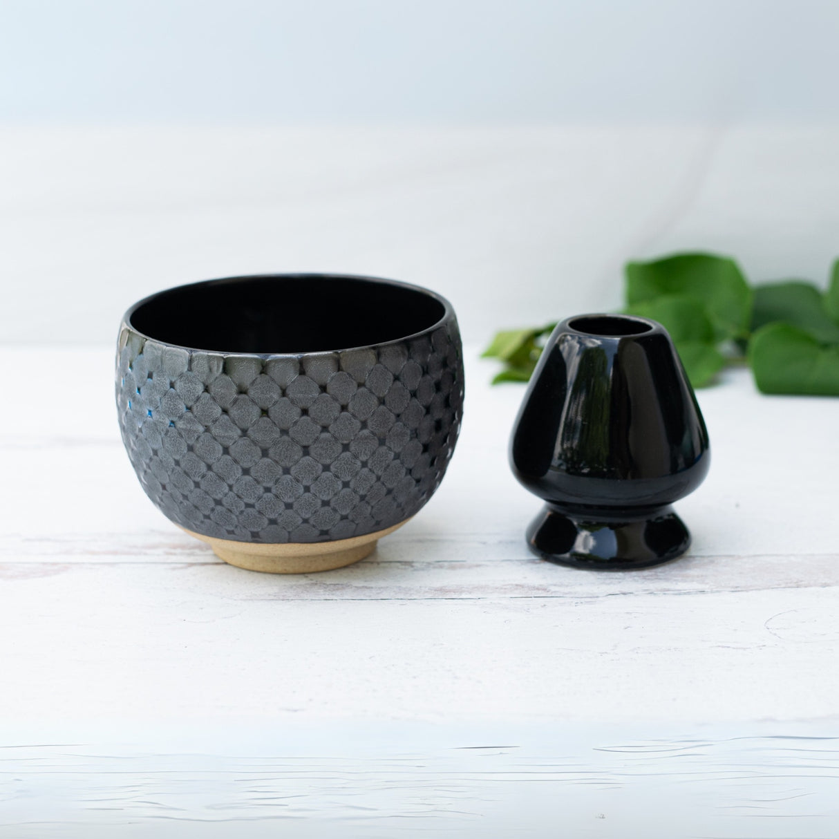 A Black Ami Pattern Matcha Bowl and a small black vase sit on a white surface, with green leaves in the background, highlighting the elegance of Japanese pottery.
