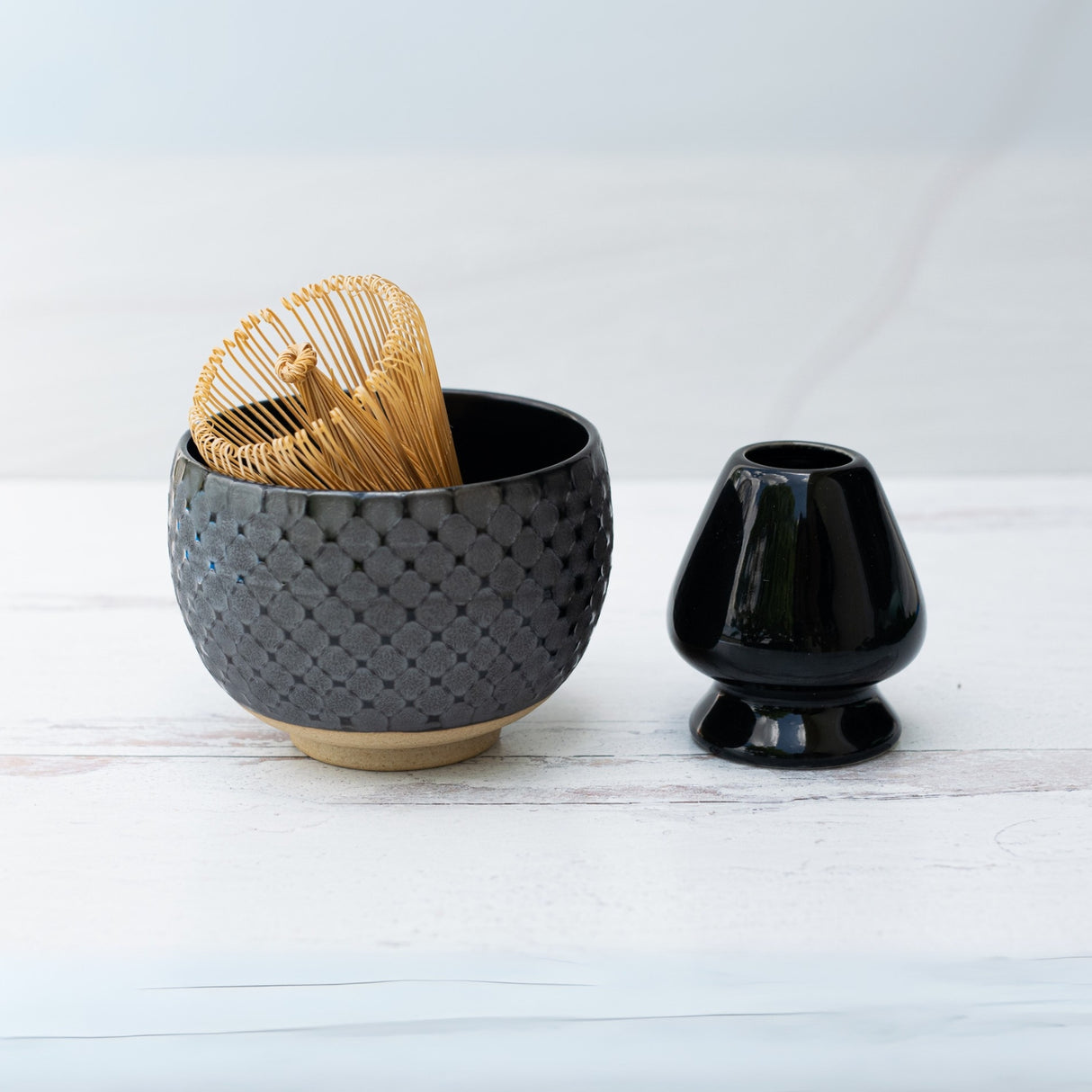 The Black Ami Pattern Matcha Bowl, along with a bamboo whisk, is elegantly arranged on a glossy black stand against a pristine white surface.