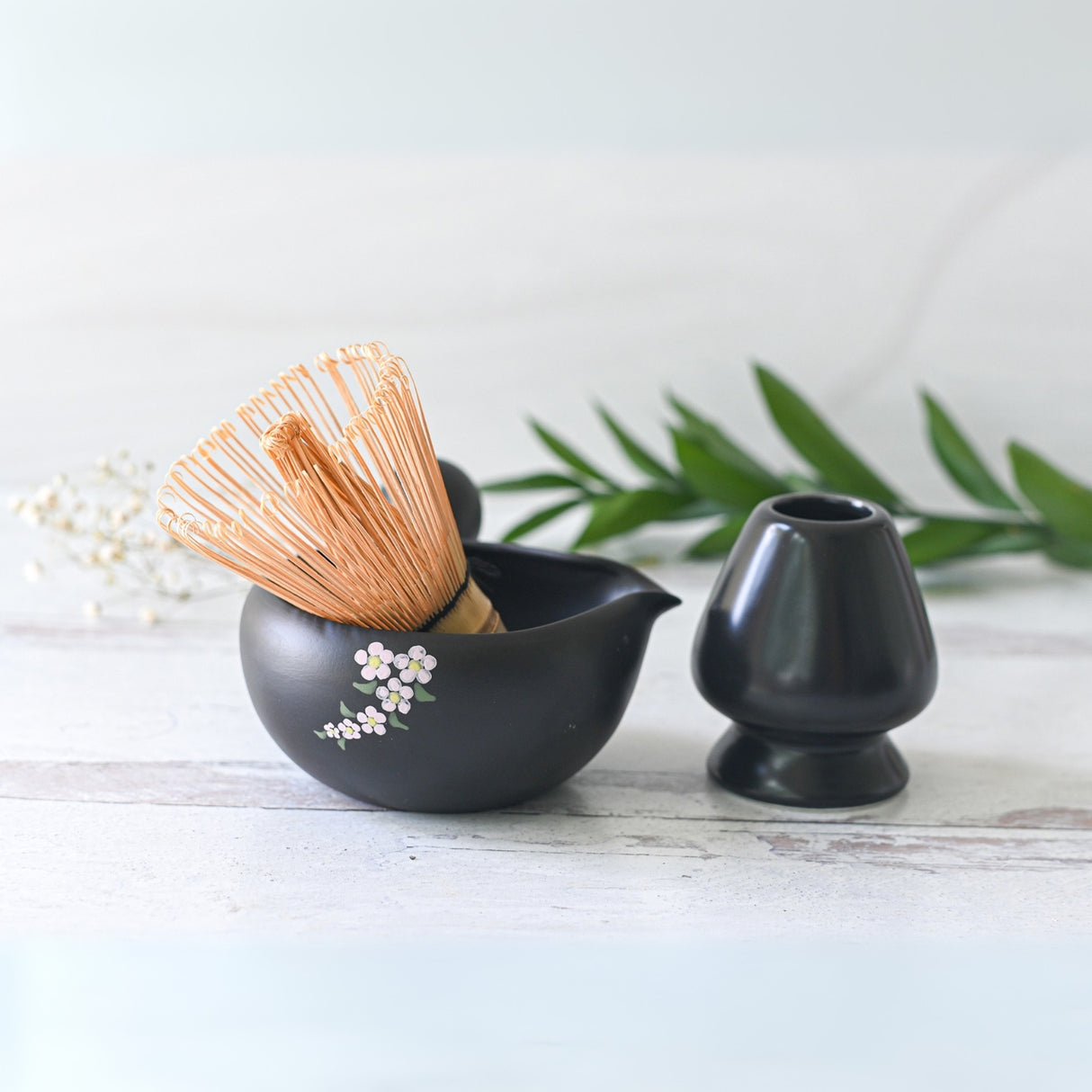 The Black Flowers Ceramic Matcha Bowl with Spout, featuring a bamboo whisk and matching stand, set against a delicate background with leaves, is perfect for enhancing Japanese tea ceremonies or as an elegant housewarming gift.