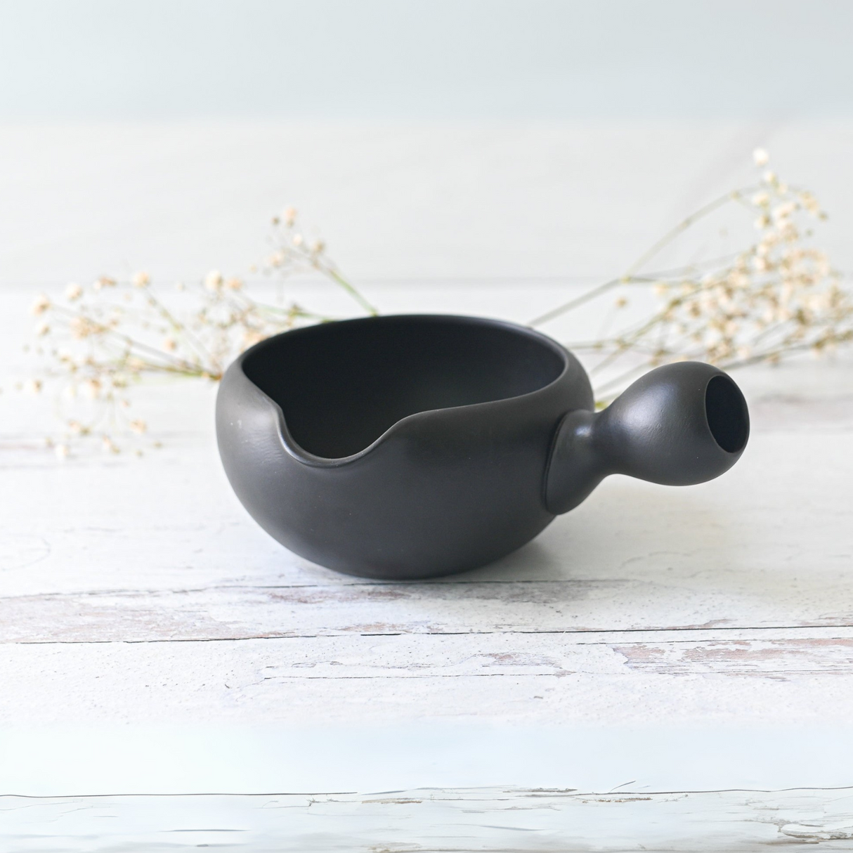 A Black Flowers Ceramic Matcha Bowl with spout rests on a light wooden surface, surrounded by delicate white flowers, making it an ideal housewarming gift.