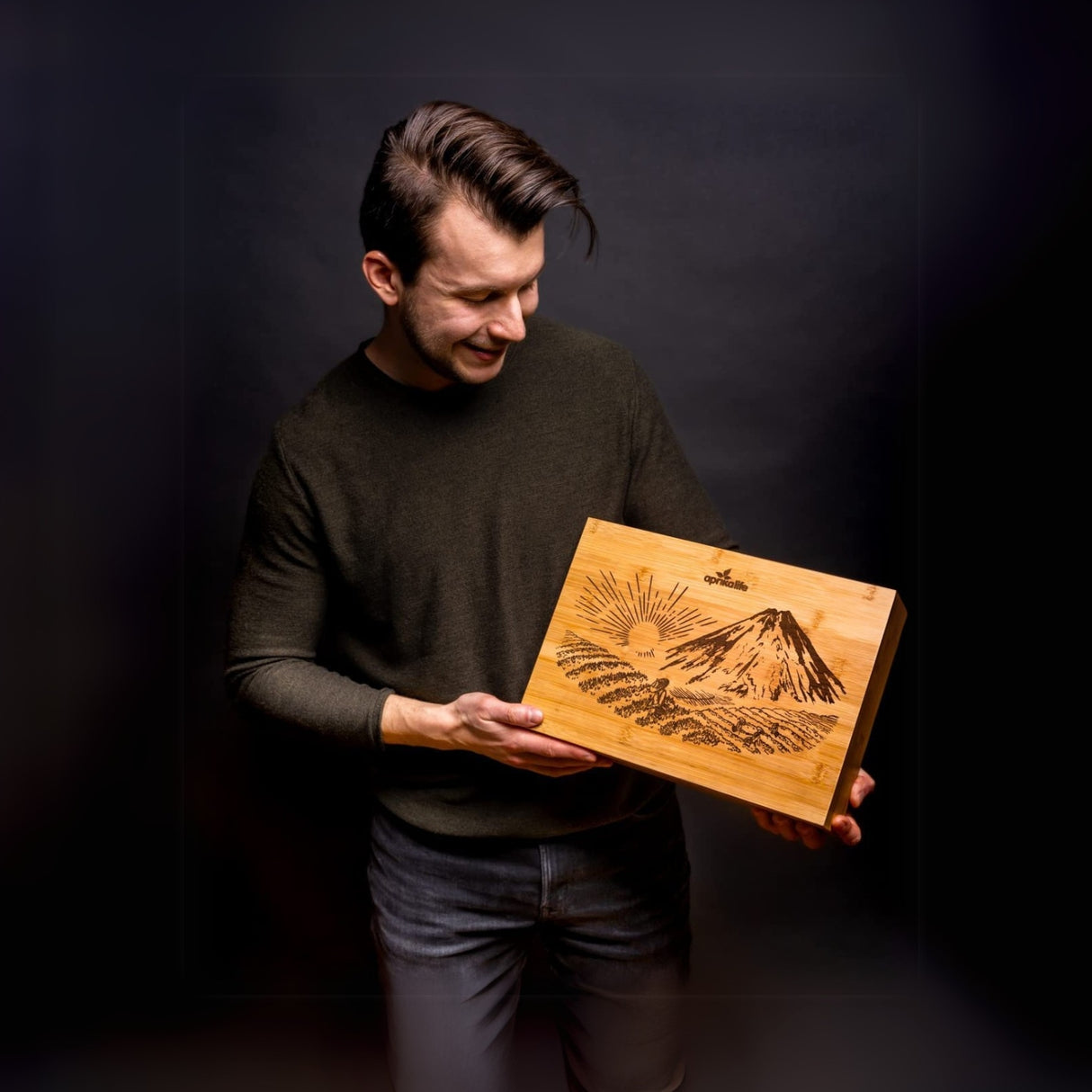 A man smiles while holding a wooden board engraved with a mountain and sun, standing against a dark backdrop, embodying the serenity of a Black Matcha Tea Gift Set ritual.