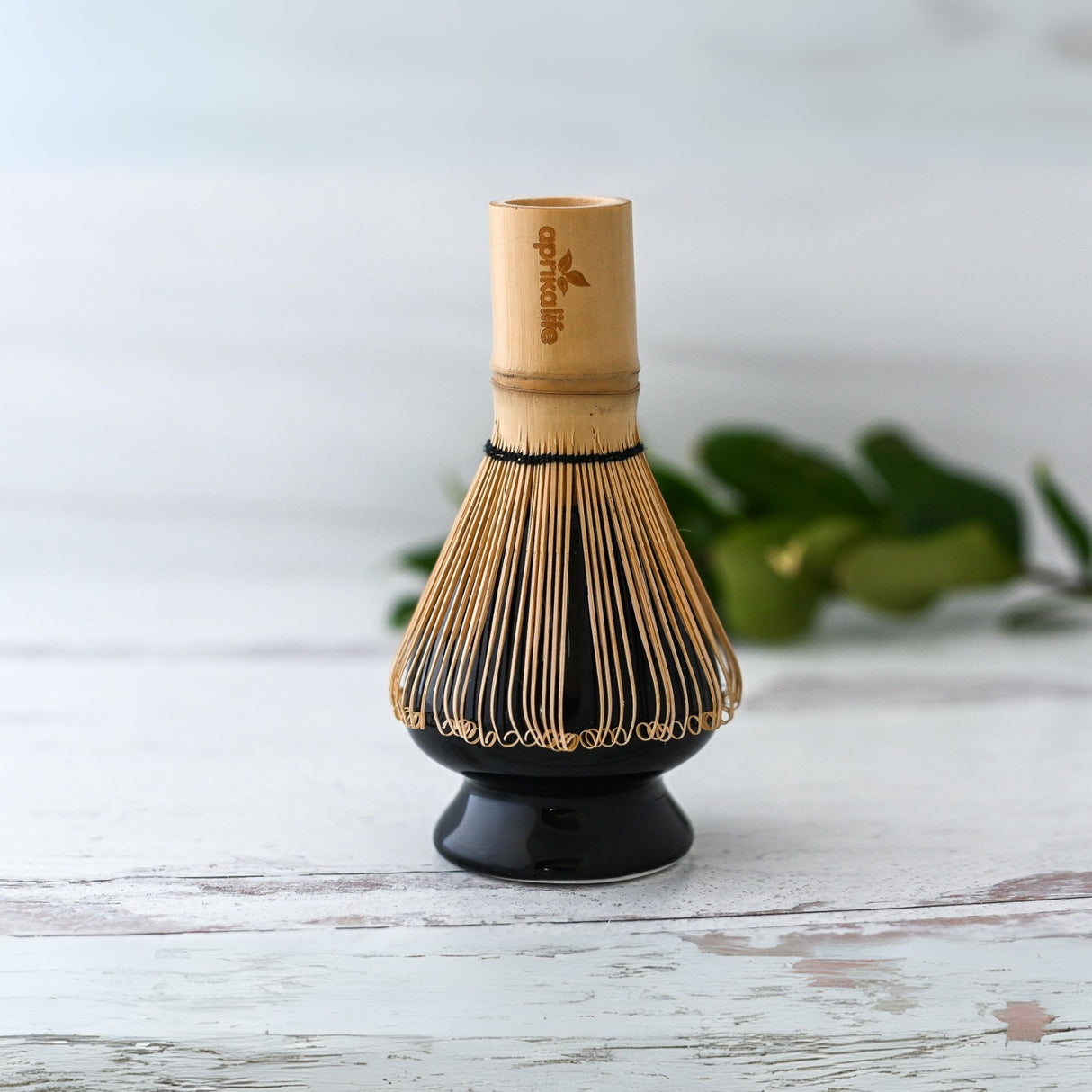 A Black Matcha Whisk Holder and Bamboo Whisk Set sits on a wooden surface, with green leaves in the background, capturing a serene Japanese aesthetic.