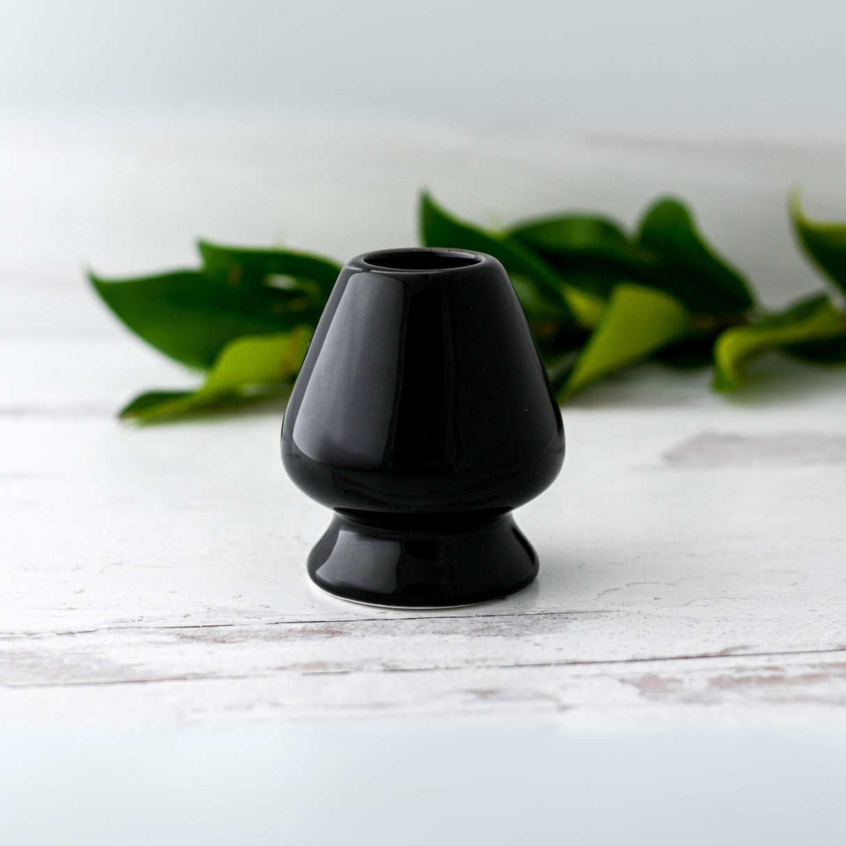 The Black Matcha Whisk Holder from the Bamboo Whisk Set sits on a white surface with green leaves in the background, evoking a serene Japanese aesthetic.