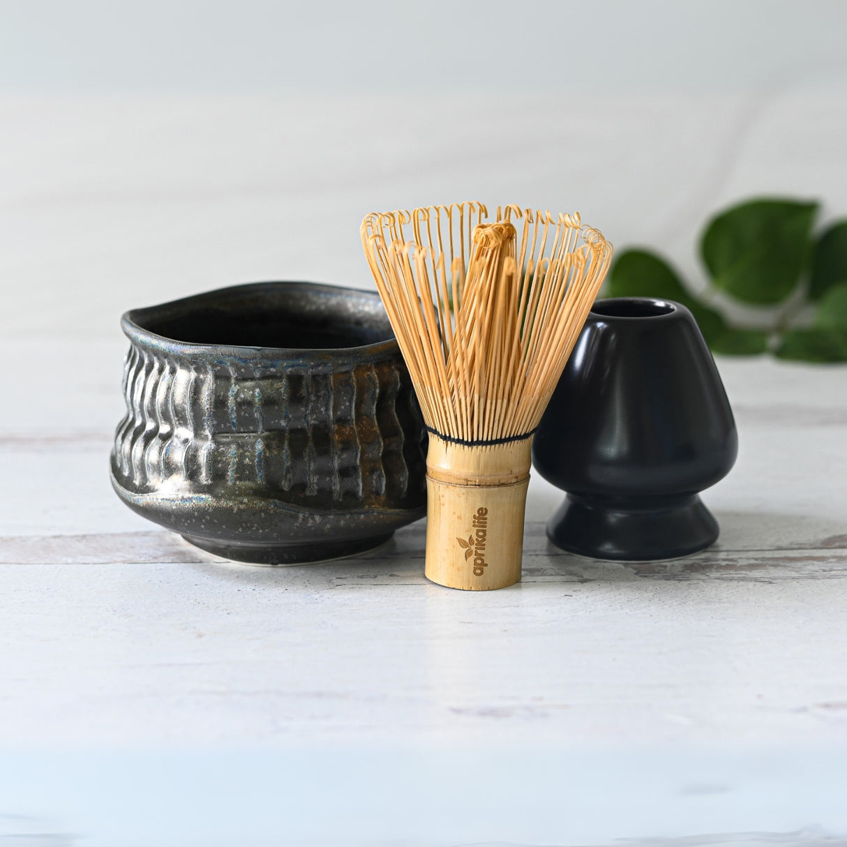 The Black Ceramic Meisen Matcha Bowl, bamboo matcha whisk, and whisk holder rest elegantly on a light wooden surface with green leaves in the background.