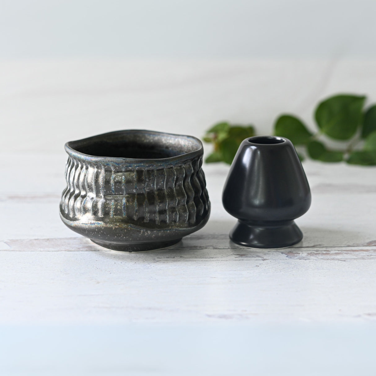 Two Black Ceramic Meisen Matcha Bowls, crafted in Minoyaki pottery, sit gracefully on a white surface with lush green leaves in the background.