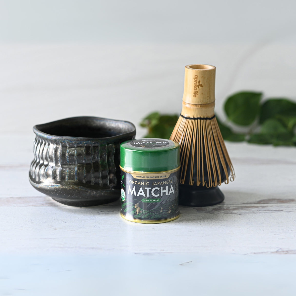 The Black Ceramic Meisen Matcha Bowl, bamboo whisk, and a tin of organic Japanese matcha are elegantly displayed on a wooden surface.