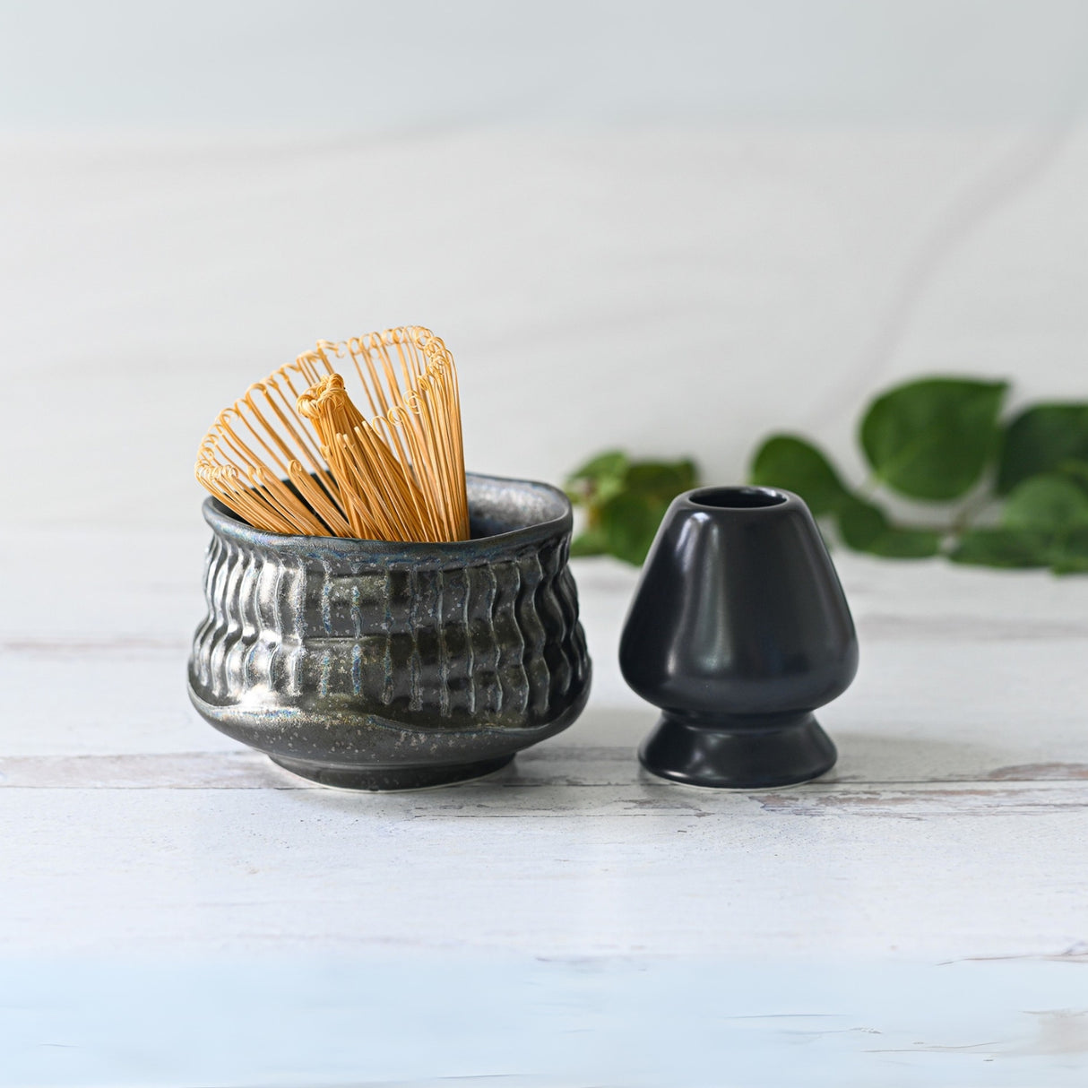 A Bamboo Matcha Whisk sits elegantly in a textured Black Ceramic Meisen Matcha Bowl, complemented by a black stand on a light surface.