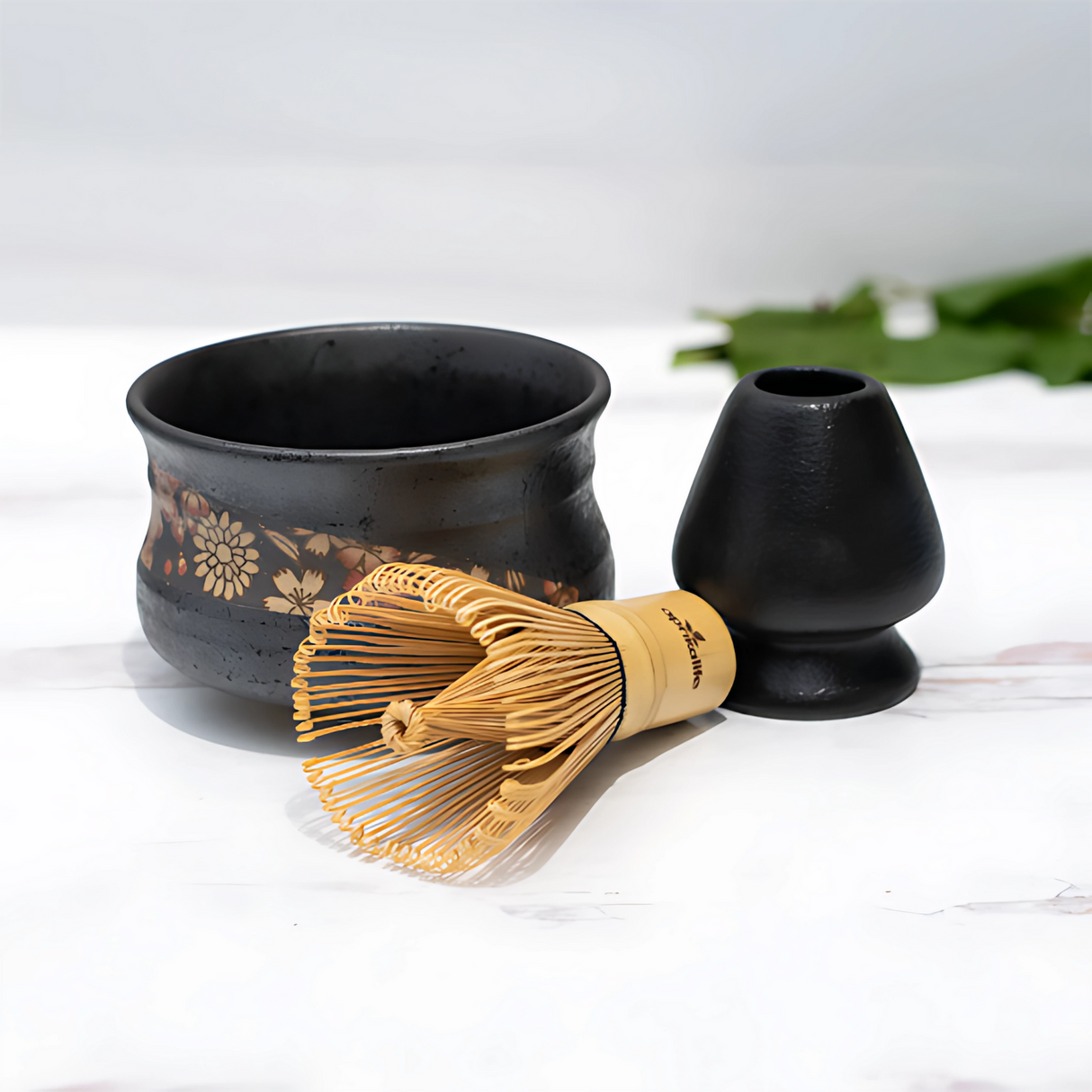 The Black with Flowers Set includes a ceramic matcha bowl, crafted using Minoyaki pottery techniques, accompanied by a bamboo matcha whisk and stand on a white surface, with a green leaf in the background.