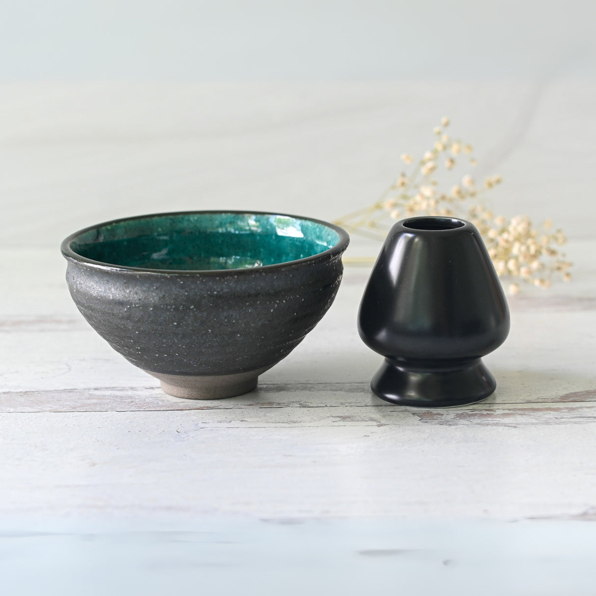 The Blue and Black Ceramic Matcha Bowl with a teal interior sits beside a small black ceramic vase on a light wooden surface.