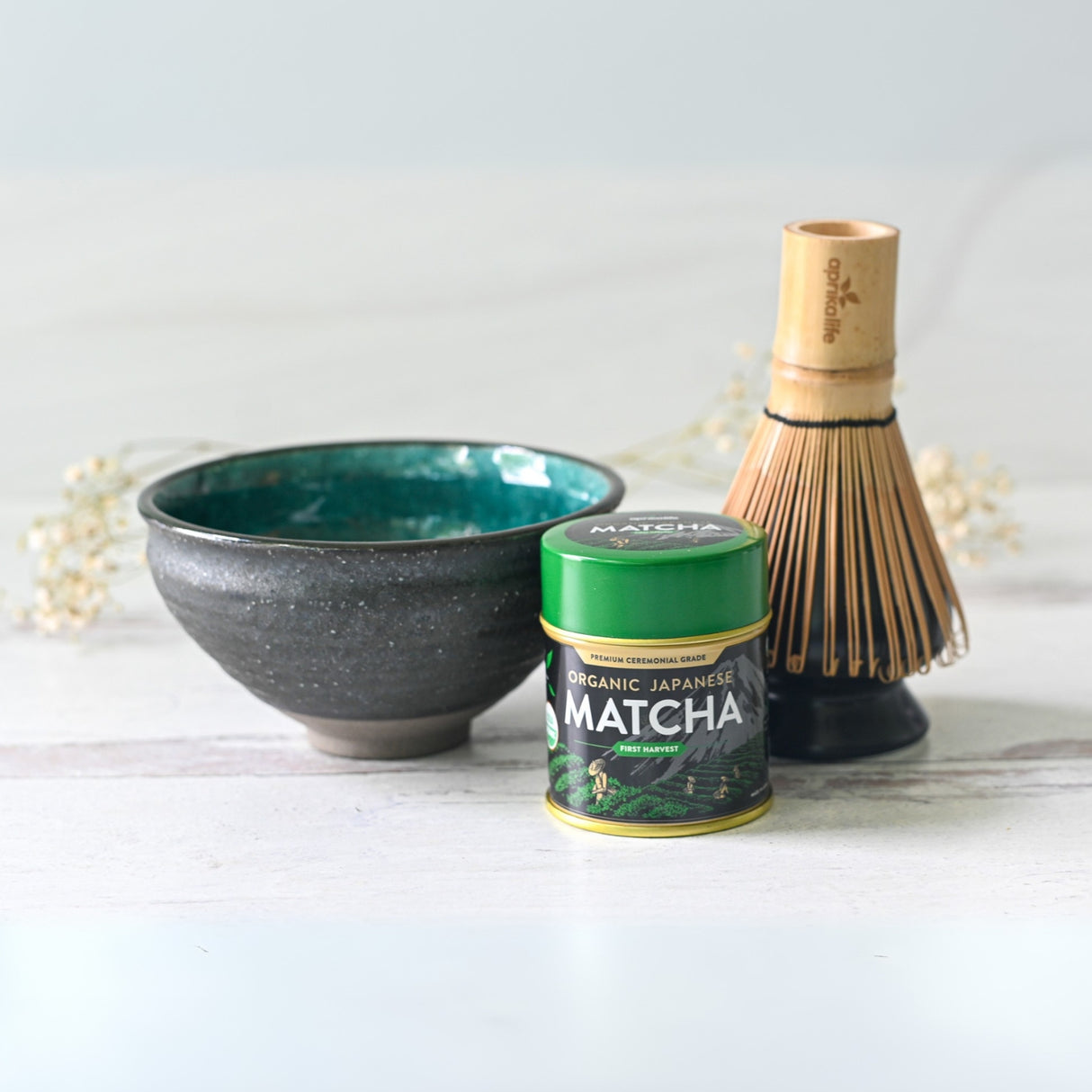 A matcha starter set showcasing a Blue and Black Ceramic Matcha Bowl, bamboo whisk, and a green can of organic Japanese matcha elegantly arranged on a white surface.