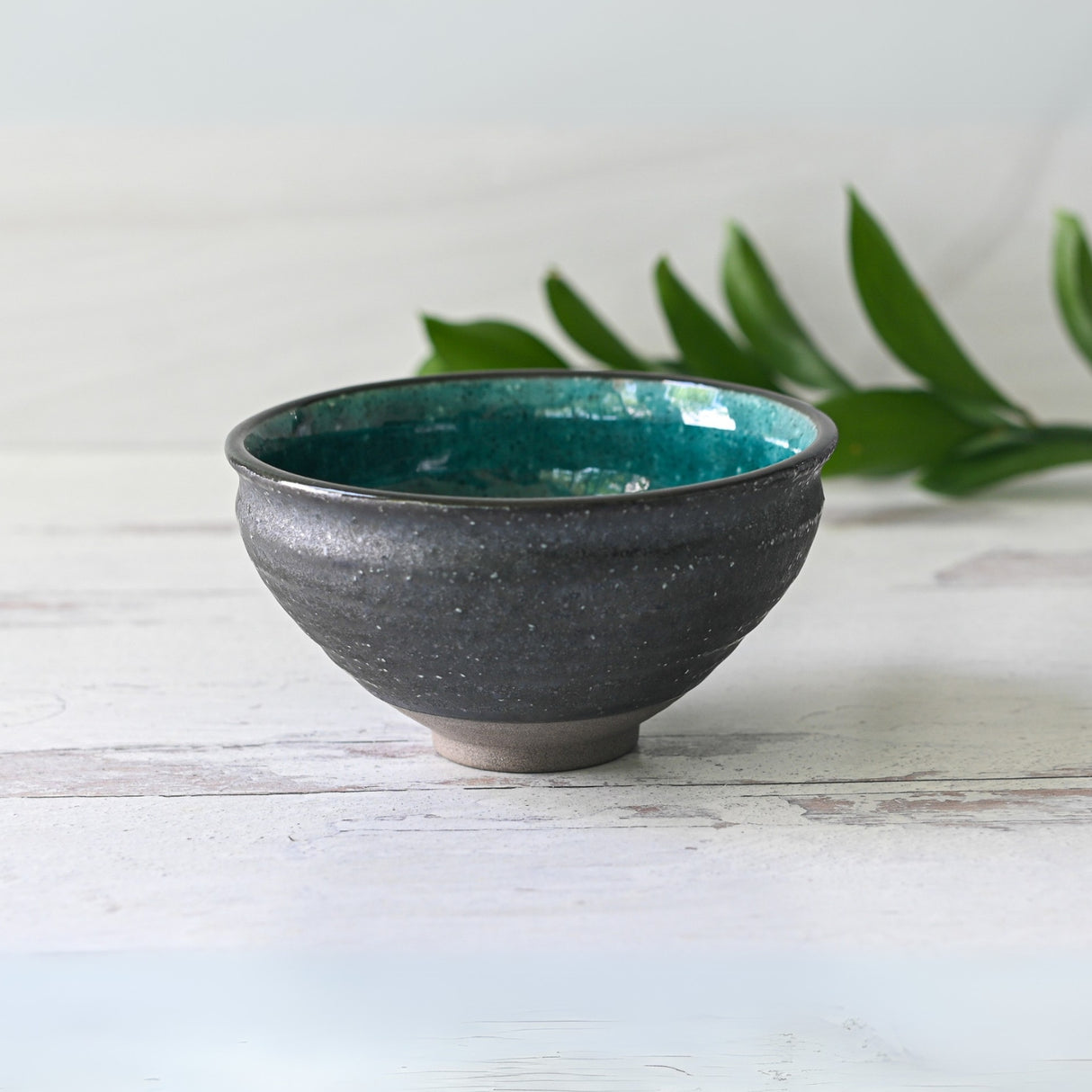 The Blue and Black Ceramic Matcha Bowl from the set, featuring a dark exterior and teal interior, rests on a light wooden surface beside green leaves. Set includes a bamboo whisk, whisk holder, and organic Japanese matcha.