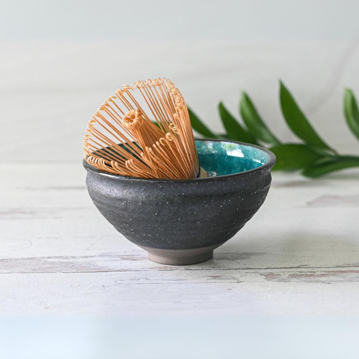 A Blue and Black Ceramic Matcha Bowl paired with a bamboo whisk, surrounded by green leaves on light wood, creates an inviting scene ideal for any matcha starter set.