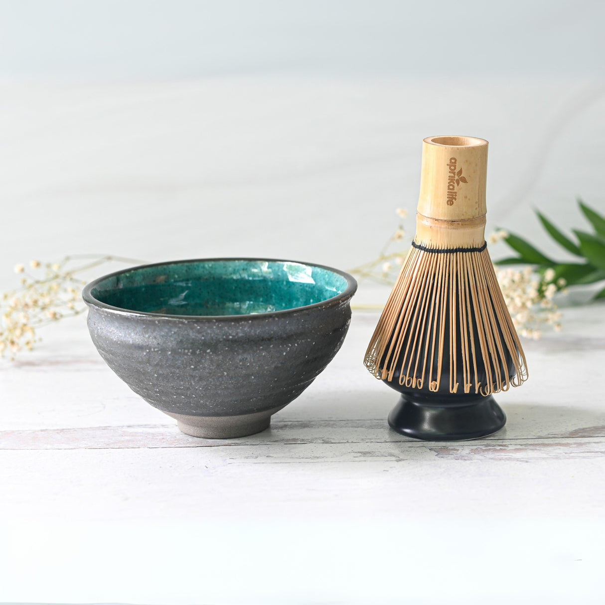 A Blue and Black Ceramic Matcha Bowl with a teal interior sits beside a bamboo whisk on a white surface, accented by decorative foliage—a perfect matcha starter set.