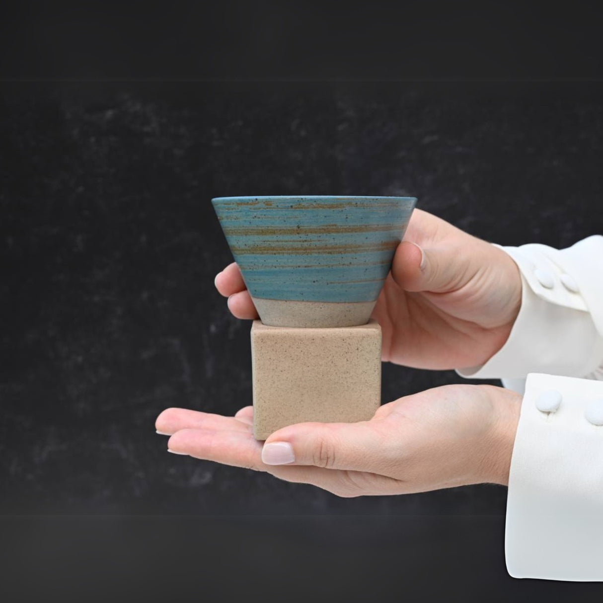 Hands hold a Blue Coffee Mug with Base - 6.8 oz/200ml, set on a stone base against a dark background.