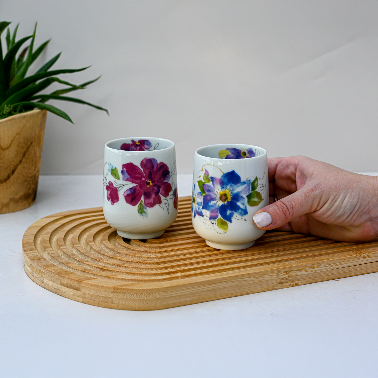 Two Blue Flower Waisted Tea Cups with floral patterns rest on a wooden tray; a hand picks up one cup, with a plant in the background.