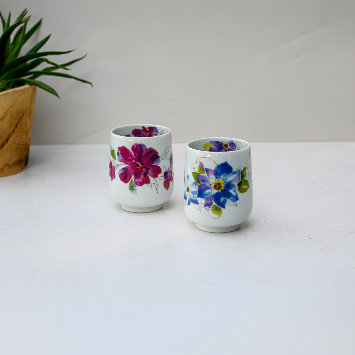 Two Blue Flower Waisted Tea Cups, featuring a handmade floral design, rest on a white surface with a potted plant in the background.
