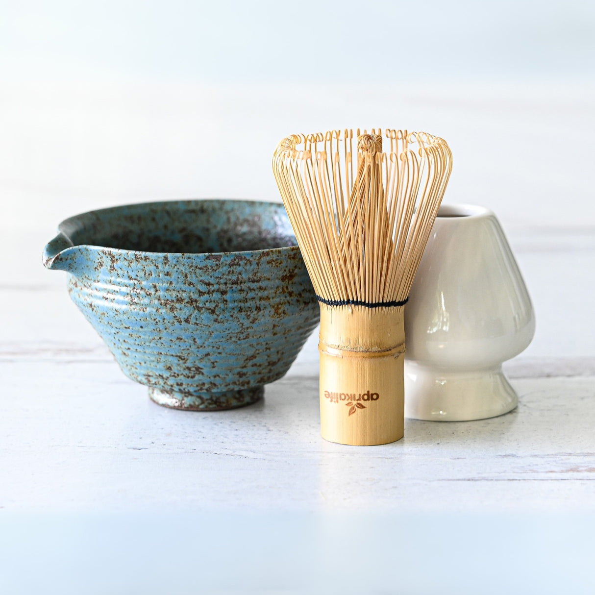 The Blue Textured Ceramic Matcha Bowl with Spout, along with a bamboo whisk and white holder, elegantly arranged on a light wooden surface—ideal for a Japanese tea ceremony or as a thoughtful housewarming gift.