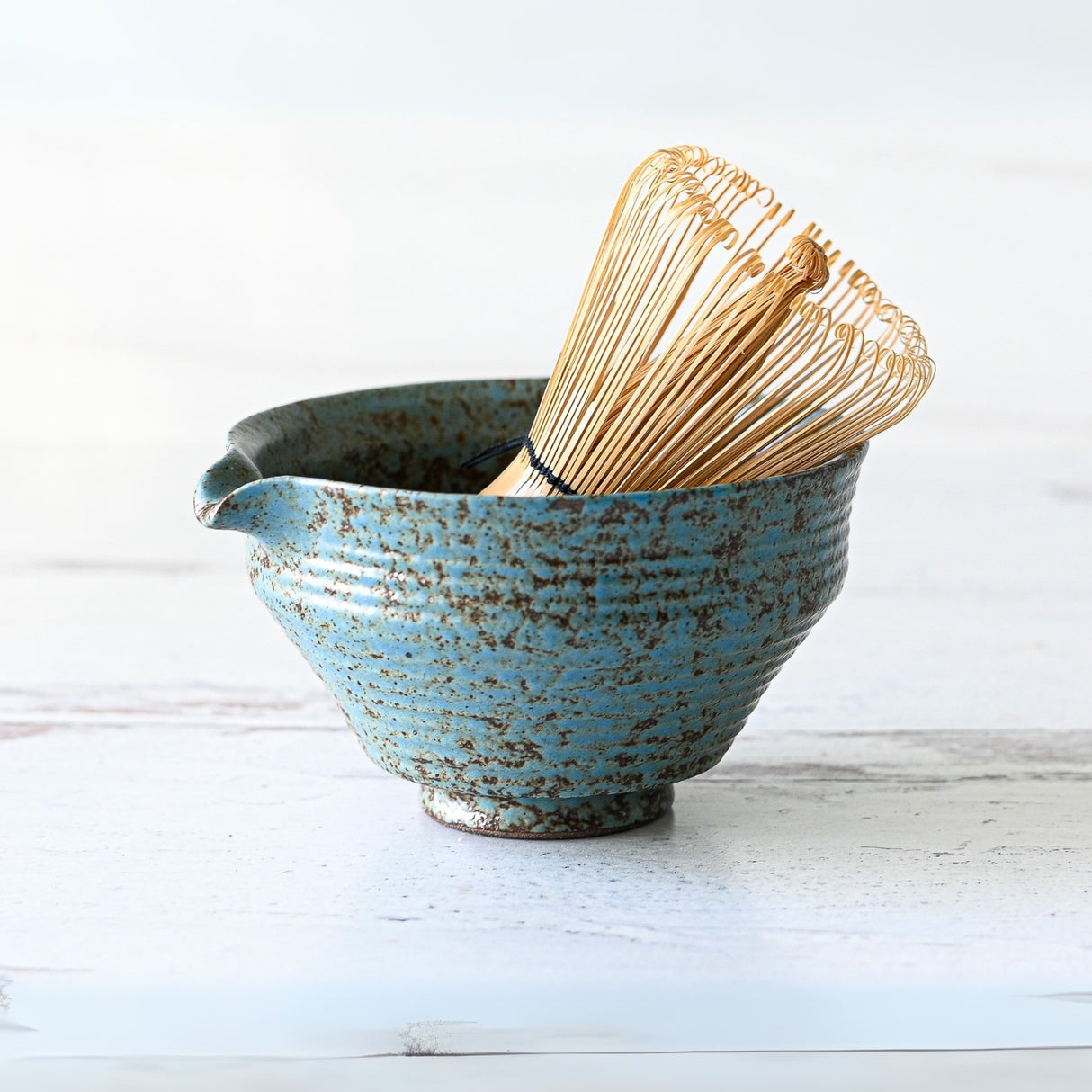 The Blue Textured Ceramic Matcha Bowl with Spout elegantly cradles a bamboo whisk on a white surface, making it perfect for Japanese tea ceremony enthusiasts or as a unique housewarming gift.