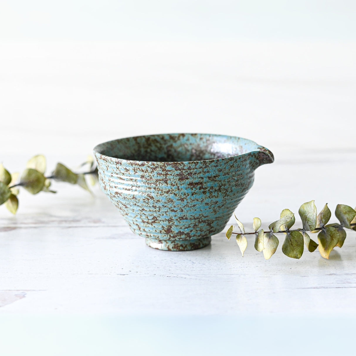 The Blue Textured Ceramic Matcha Bowl with Spout, paired with a bamboo matcha whisk and holder, rests on a white surface, surrounded by eucalyptus for a tranquil Japanese tea ceremony vibe.