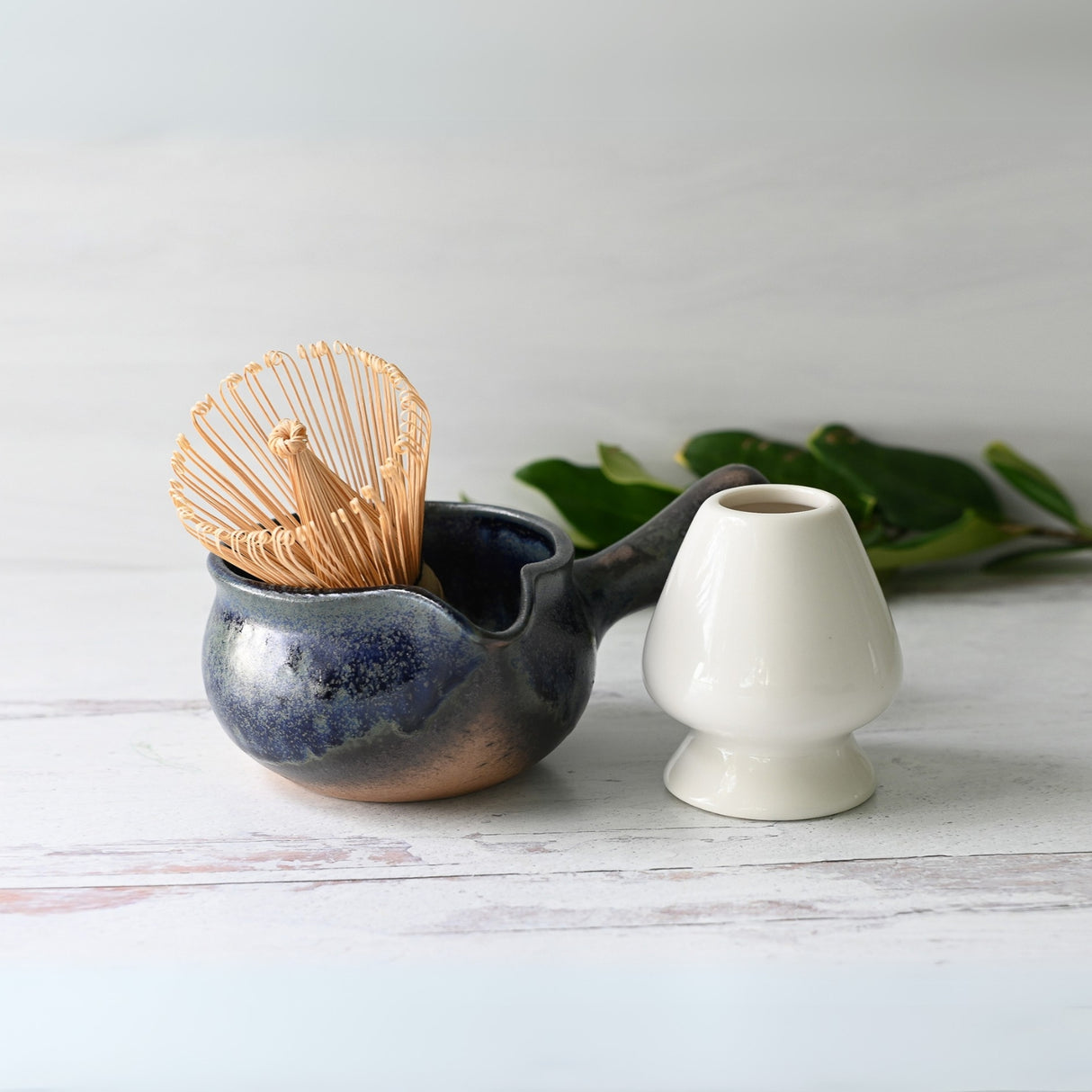 The Blue Spouted Ceramic Matcha Bowl and Bamboo Matcha Whisk from the Organic Japanese Matcha Set are placed next to a white vase on a light surface, with green leaves in the background.