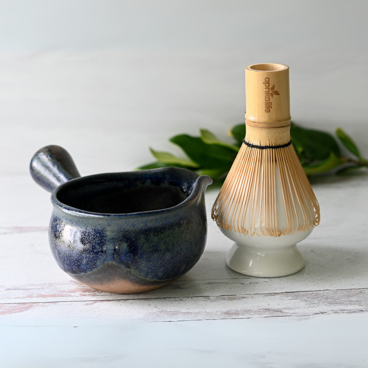 A Blue Spouted Ceramic Matcha Bowl paired with a Bamboo Matcha Whisk rests on a light surface, complemented by green leaves in the background.