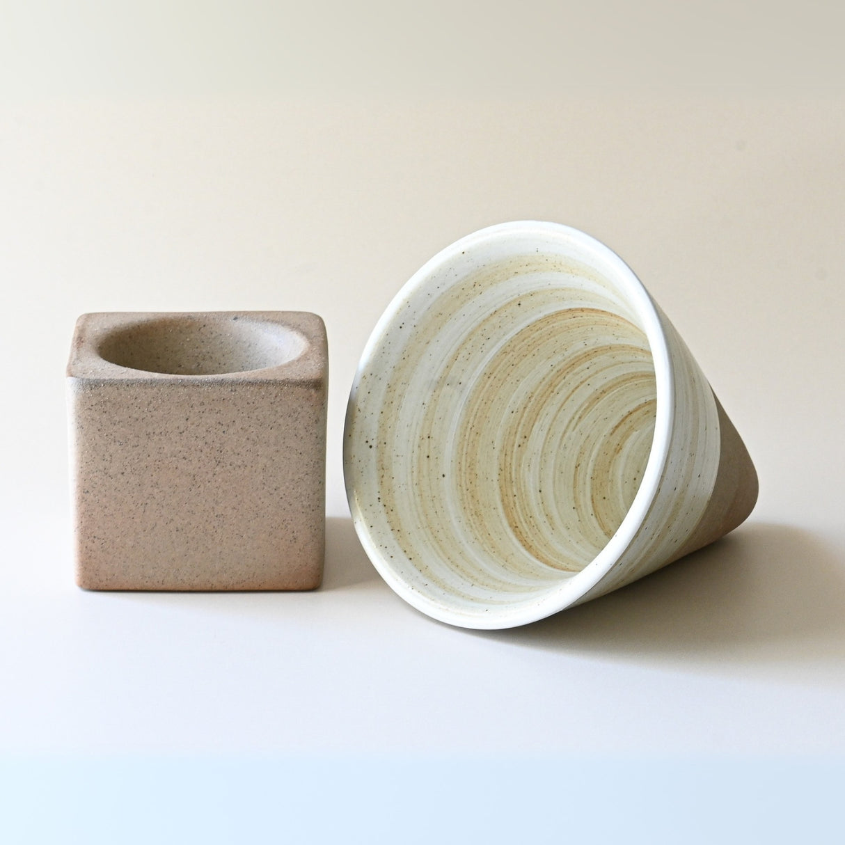 A square and a round ceramic vase, one upright and another on its side, rest elegantly on a plain surface, reminiscent of the grace of a Beige Coffee Mug with Base - 6.8 oz/200ml waiting to be admired.