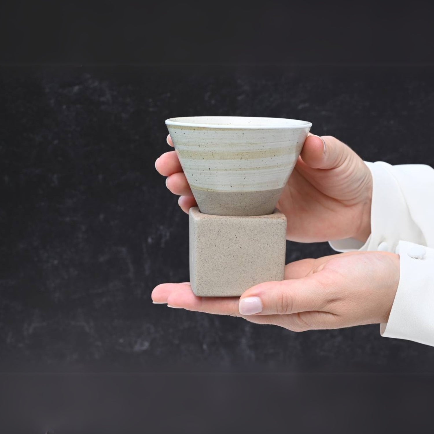 Hands holding a Beige Coffee Mug with Base - 6.8 oz/200ml, set against a dark background.
