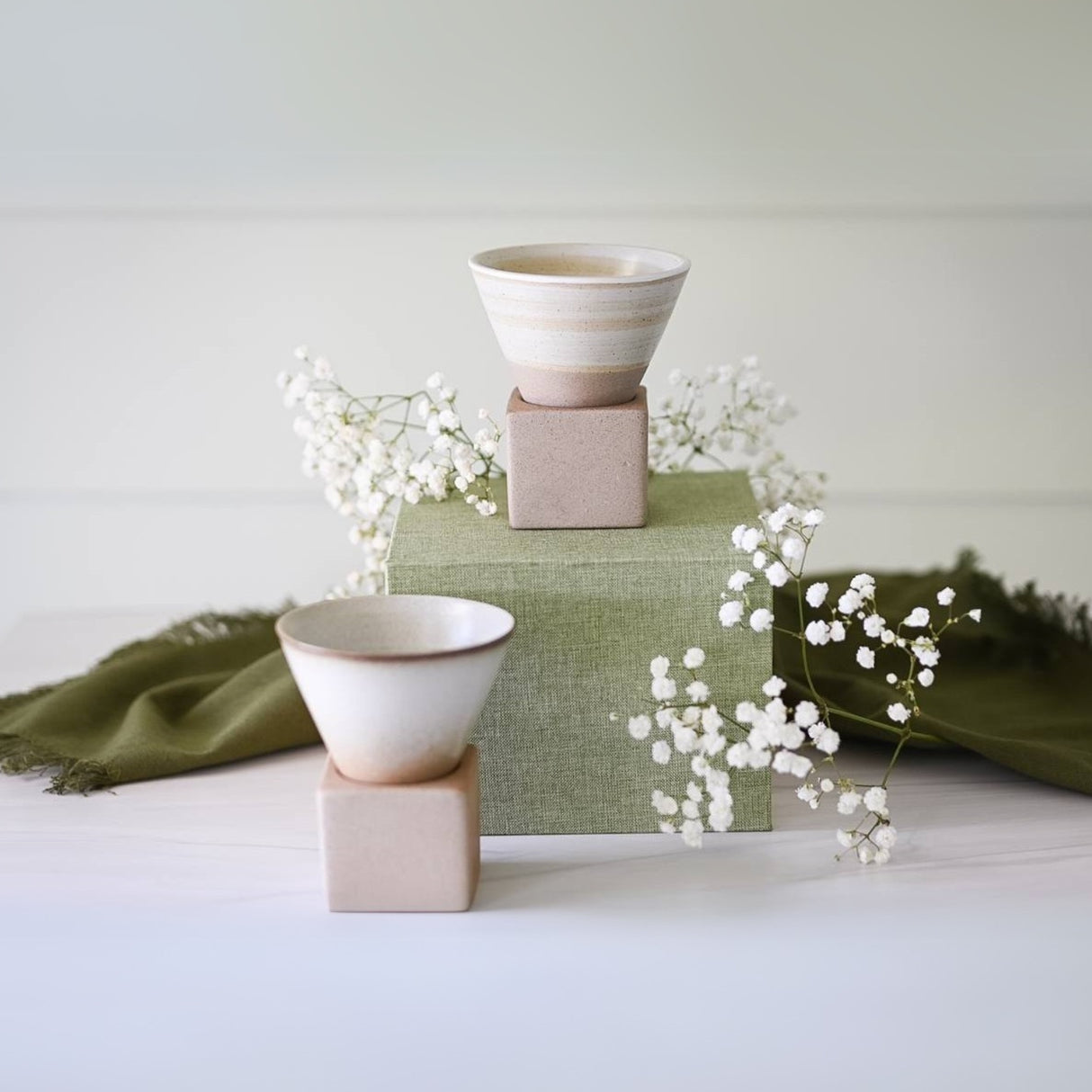 Two Beige Coffee Mugs with 6.8 oz bases, surrounded by white flowers and a green cloth on a white surface, create an enchanting scene perfect for any setting.