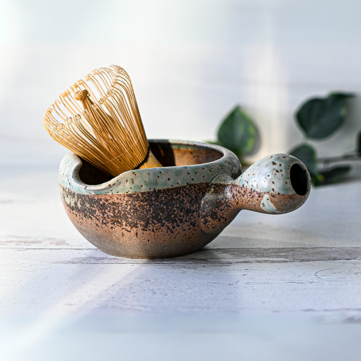 The Brown Ceramic Matcha Bowl with Spout, Bamboo Whisk, Whisk Holder and Organic Japanese Matcha Set sits on a light surface, evoking the calm of a tea ceremony amid blurred green leaves in the background.