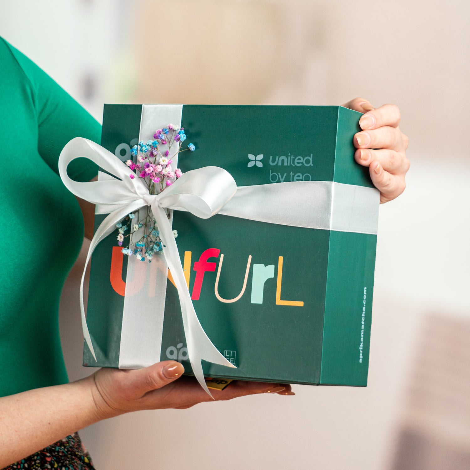 A person holds a blue "Unfurl" Ceremonial Matcha Starter Kit, wrapped with white ribbon and flowers—the perfect gift for those inspired by Japanese tea ceremonies.