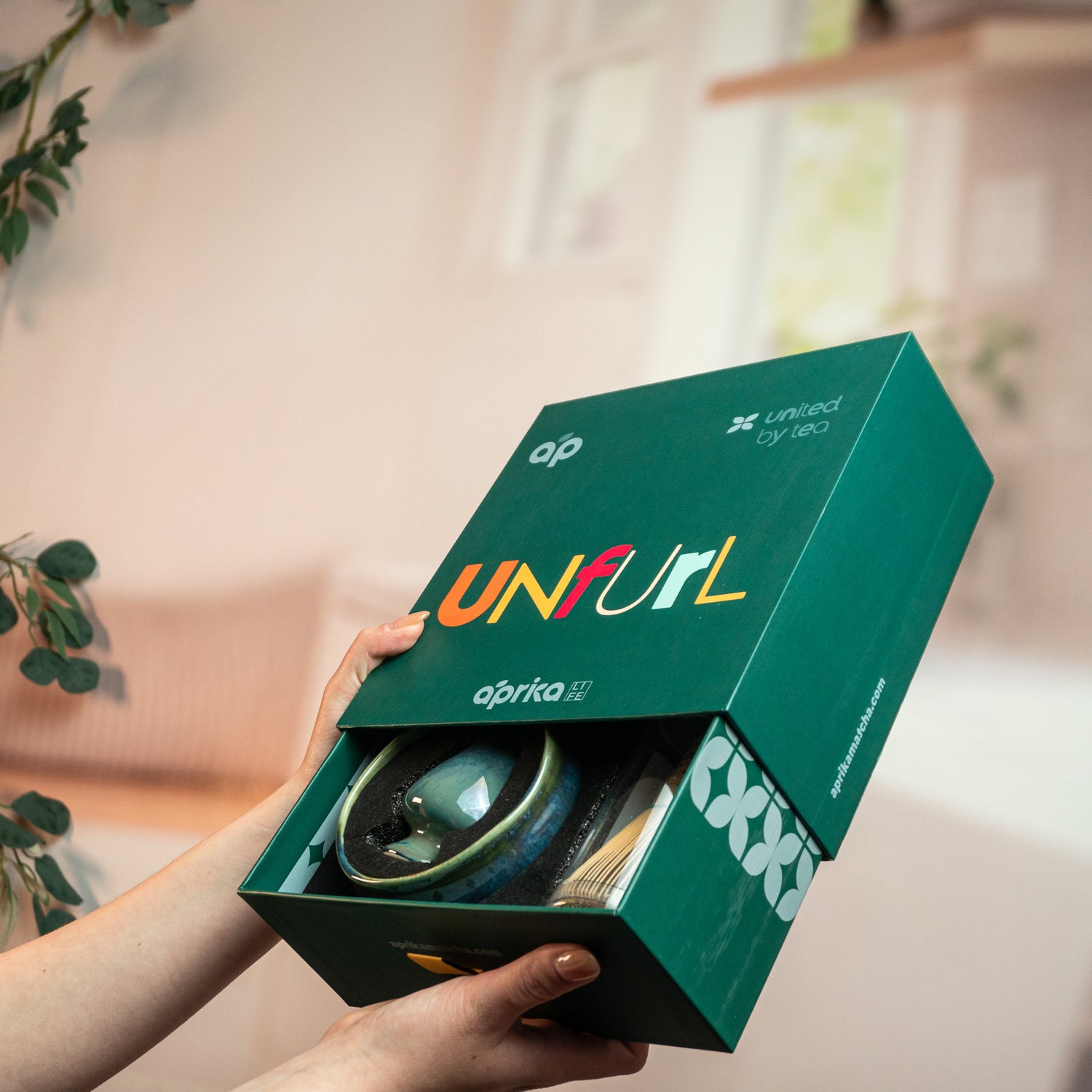 Hands holding a blue box labeled "UNFURL," part of the Ceremonial Matcha Starter Kit, with a round object and plants in the background—evoking the spirit of Japanese tea ceremonies.