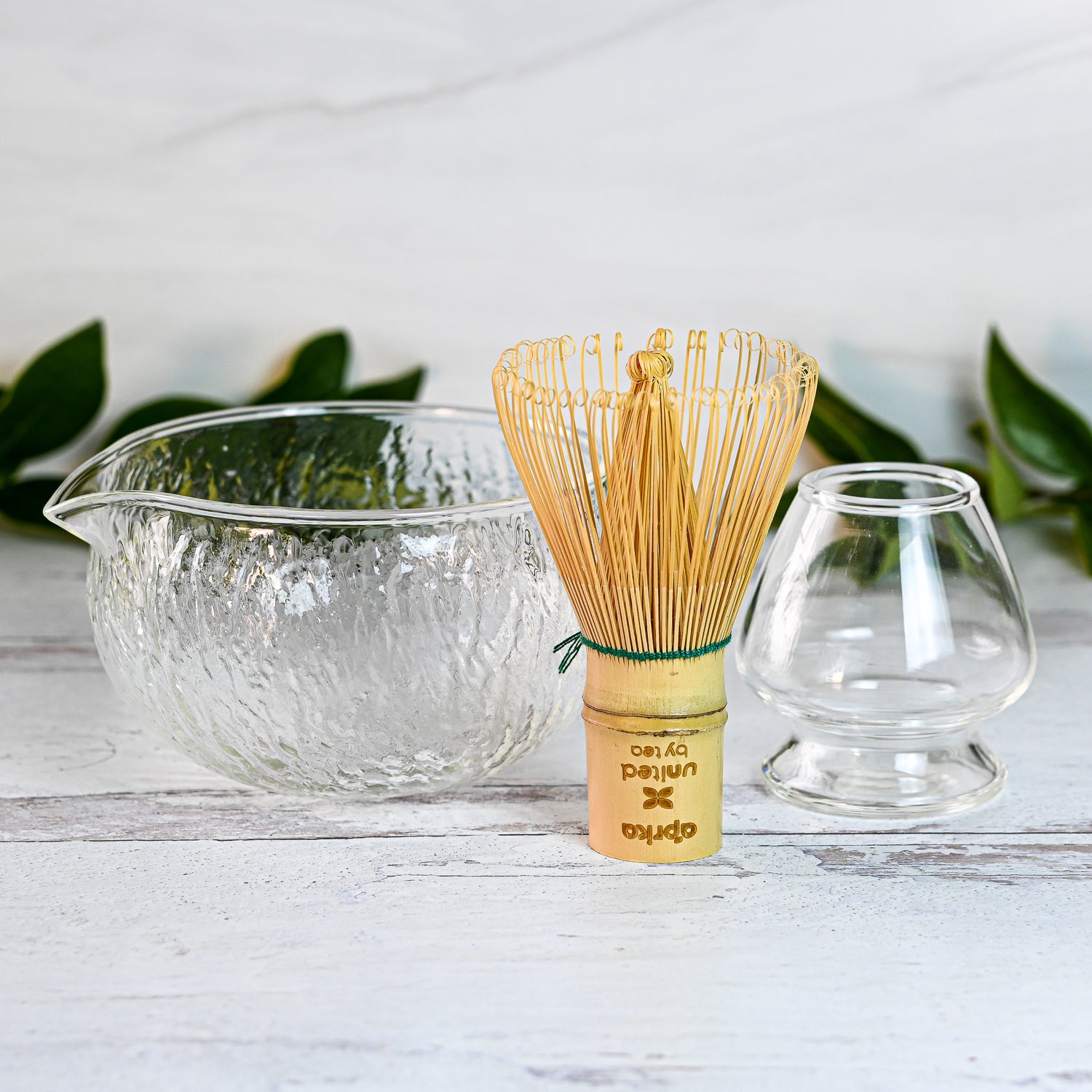 The Textured Spouted Glass Matcha Bowl Pattern 2, a bamboo matcha whisk, and a small glass cup rest elegantly on a white surface with green leaves providing a serene backdrop.