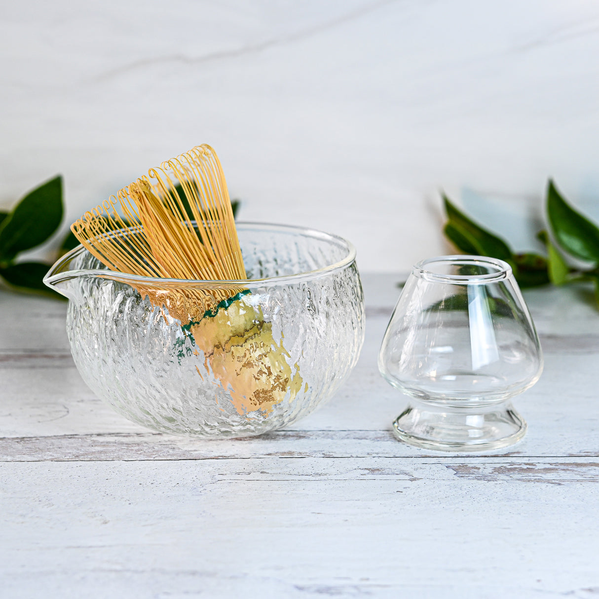 The Textured Spouted Glass Matcha Bowl Pattern 2 rests next to a Bamboo Matcha Whisk and Whisk Holder Set on a white wooden surface, gently framed by leaves.