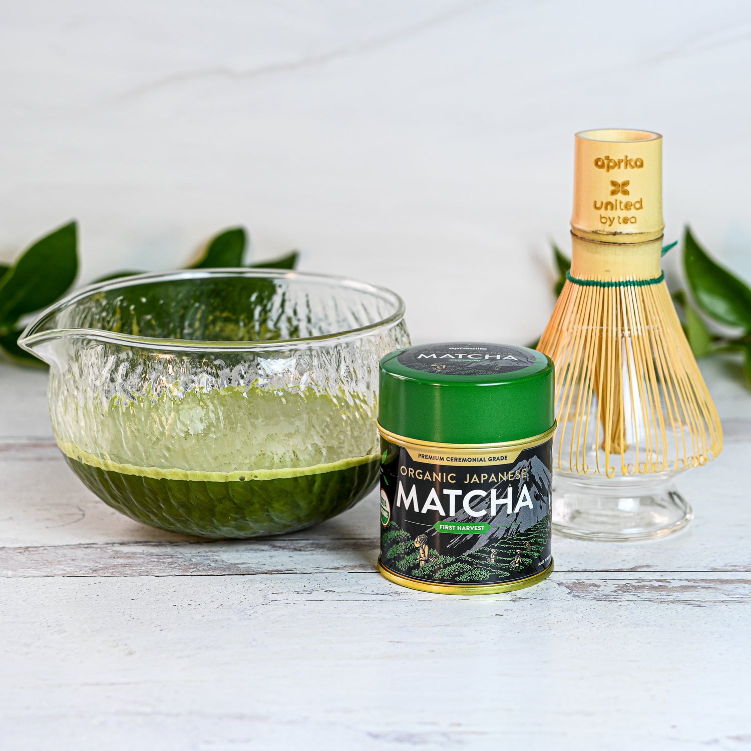 The Textured Spouted Glass Matcha Bowl Pattern 1, paired with matcha tea, an organic matcha tin, and a bamboo whisk on a white wooden surface, is ideal for elevating your matcha ritual or serving as a sophisticated housewarming gift.