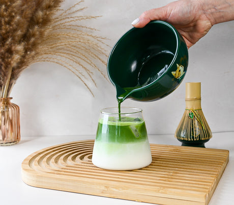 A hand pours matcha tea from the Green Unfurl Matcha Gift Set into a glass of milk on a wooden tray, with decorative items and the traditional set in the background.