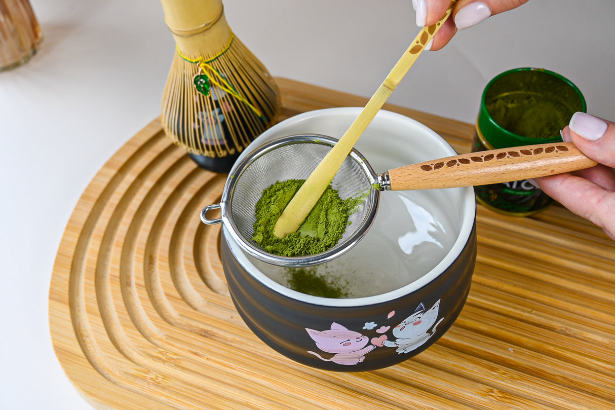 A hand sifts matcha powder into a kawaii cat bowl from the Black Unfurl Matcha Gift Set, arranged on a wooden tray with a bamboo whisk nearby.