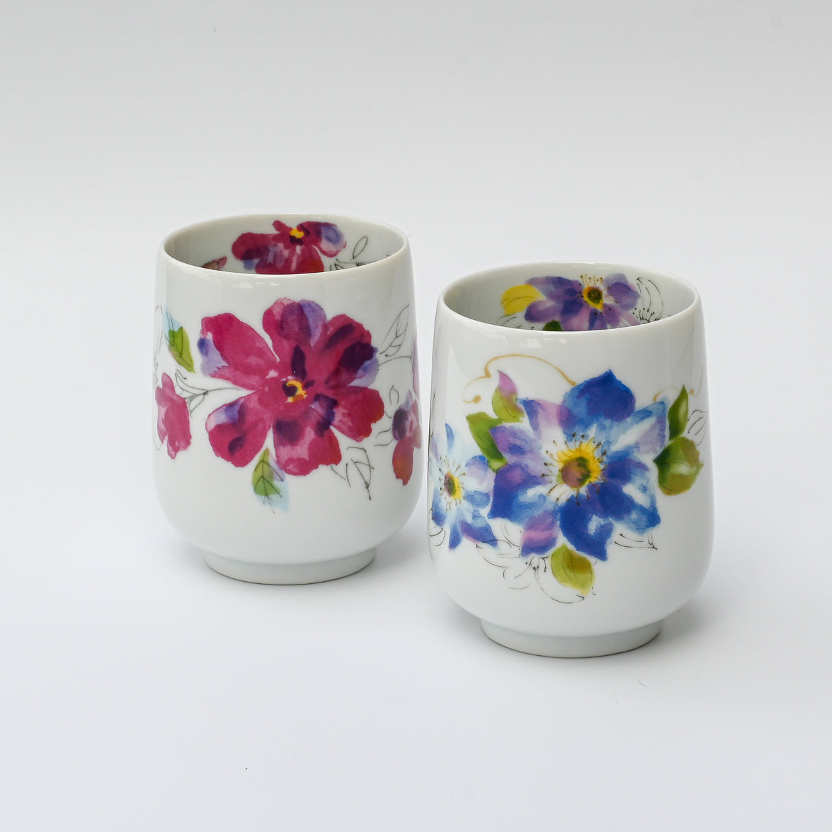 Two white Pink Flower Waisted Tea Cups, crafted using traditional Japanese ceramic techniques—one with red flowers, the other with blue and yellow blooms—are exquisite handmade art pieces.